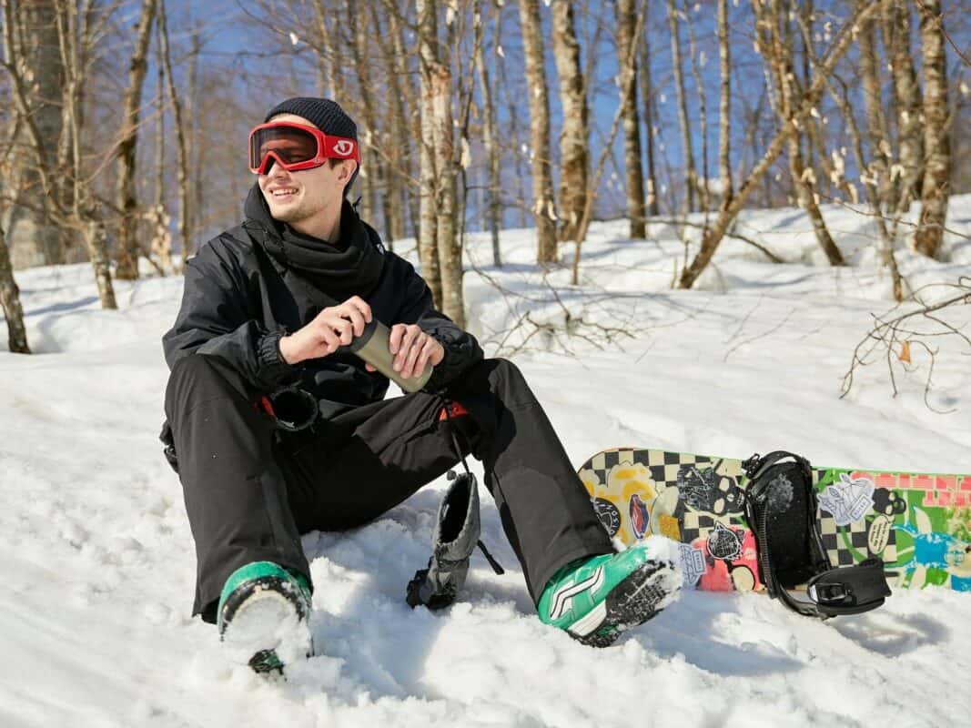 Young-male-snowboarder-wearing-black-snow-gear-sitting-in-snow-next-to-his-snowboard-for-a-water-break